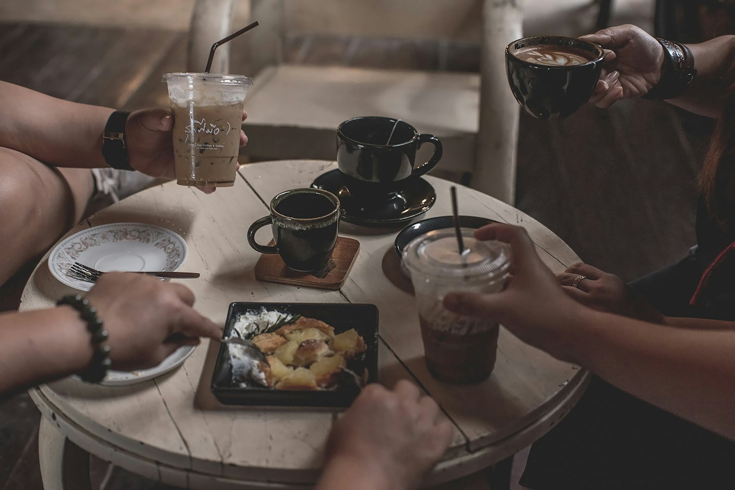 Vrienden delen koffie en dessert rond een houten cafétafel.